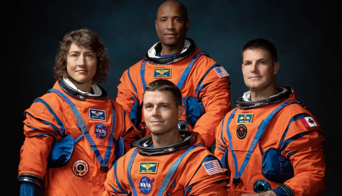 Cztery osoby w pomarańczowych skafandrach kosmicznych z widocznymi naszywkami NASA oraz flagami państw, ustawione w szeregu na neutralnym, ciemnym tle, tworzą oficjalny portret załogi.