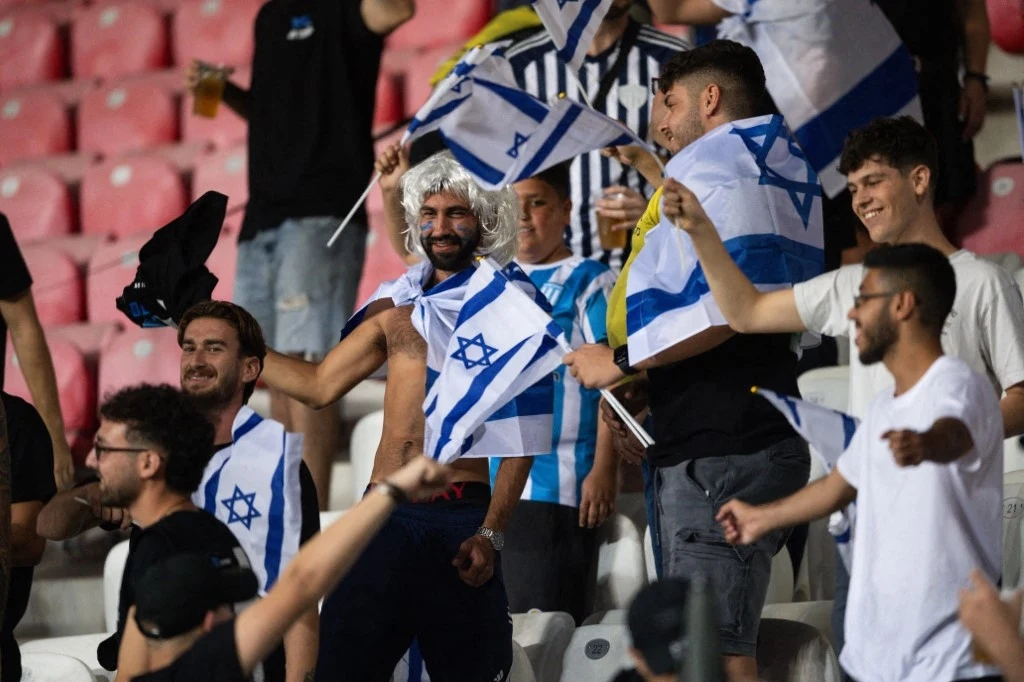 Grupa uśmiechniętych kibiców machających flagami Izraela na trybunach stadionu sportowego, część z nich ubrana w koszulki i sportowe akcesoria, w tle widoczne czerwone krzesełka.