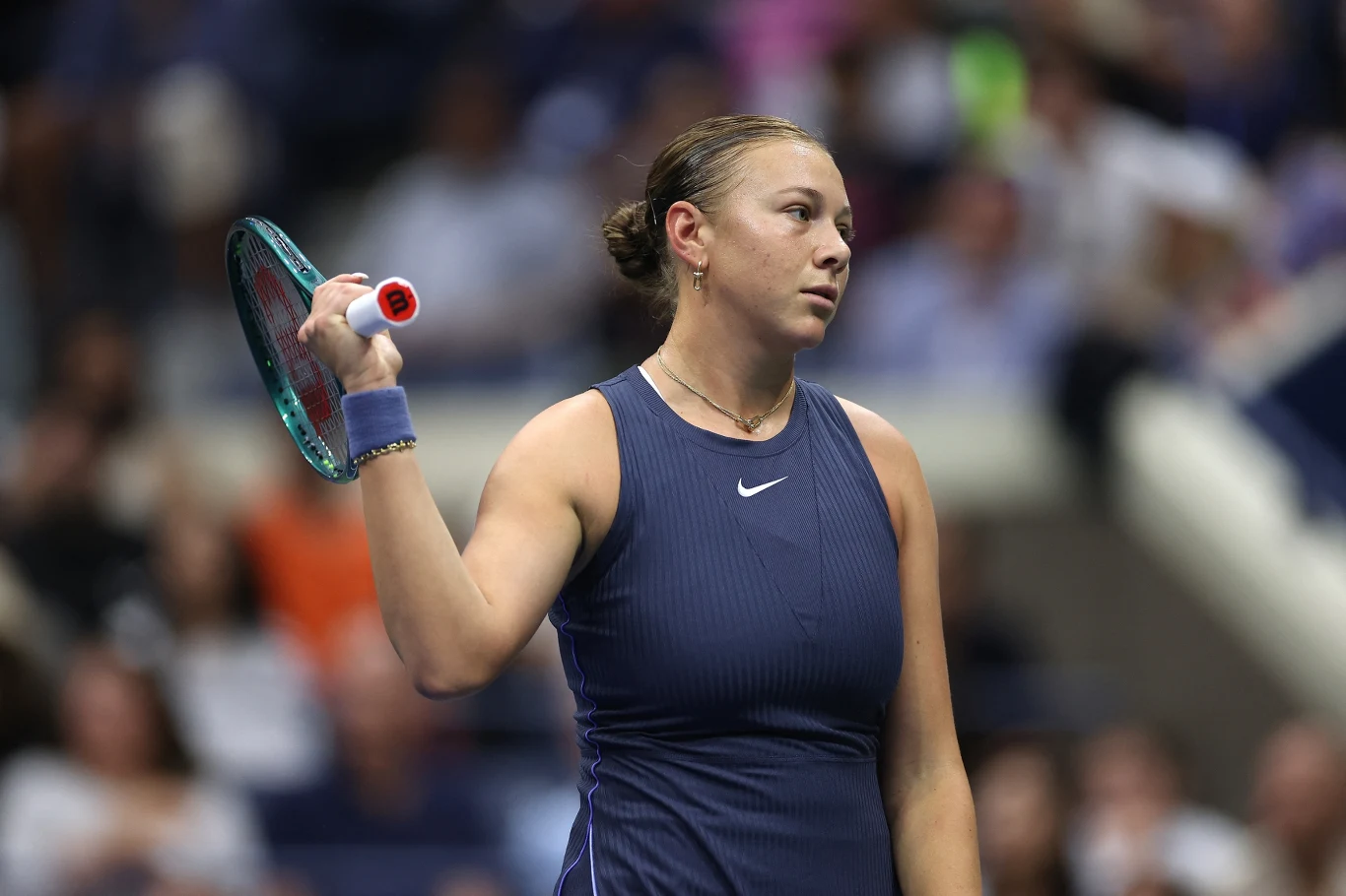 Tenisistka w granatowym stroju trzymająca rakietę tenisową w jednej ręce w trakcie meczu, wyrażająca emocje twarzą, w tle rozmyta widownia stadionu.