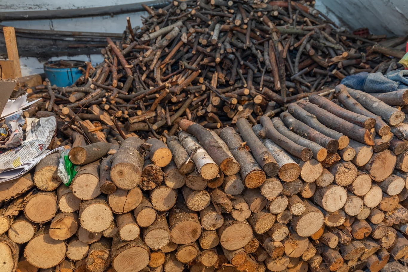 Ułożone i poukładane kawałki drewna opałowego tworzące stos, w tle widoczna większa ilość drobnych gałęzi i kawałków drewna.