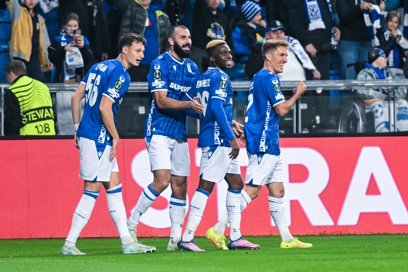 Czterech piłkarzy drużyny w niebieskich koszulkach cieszy się na boisku, w tle tłum kibiców i widoczne barwy klubowe, piłkarze kierują się razem w stronę aparatu w atmosferze radości.