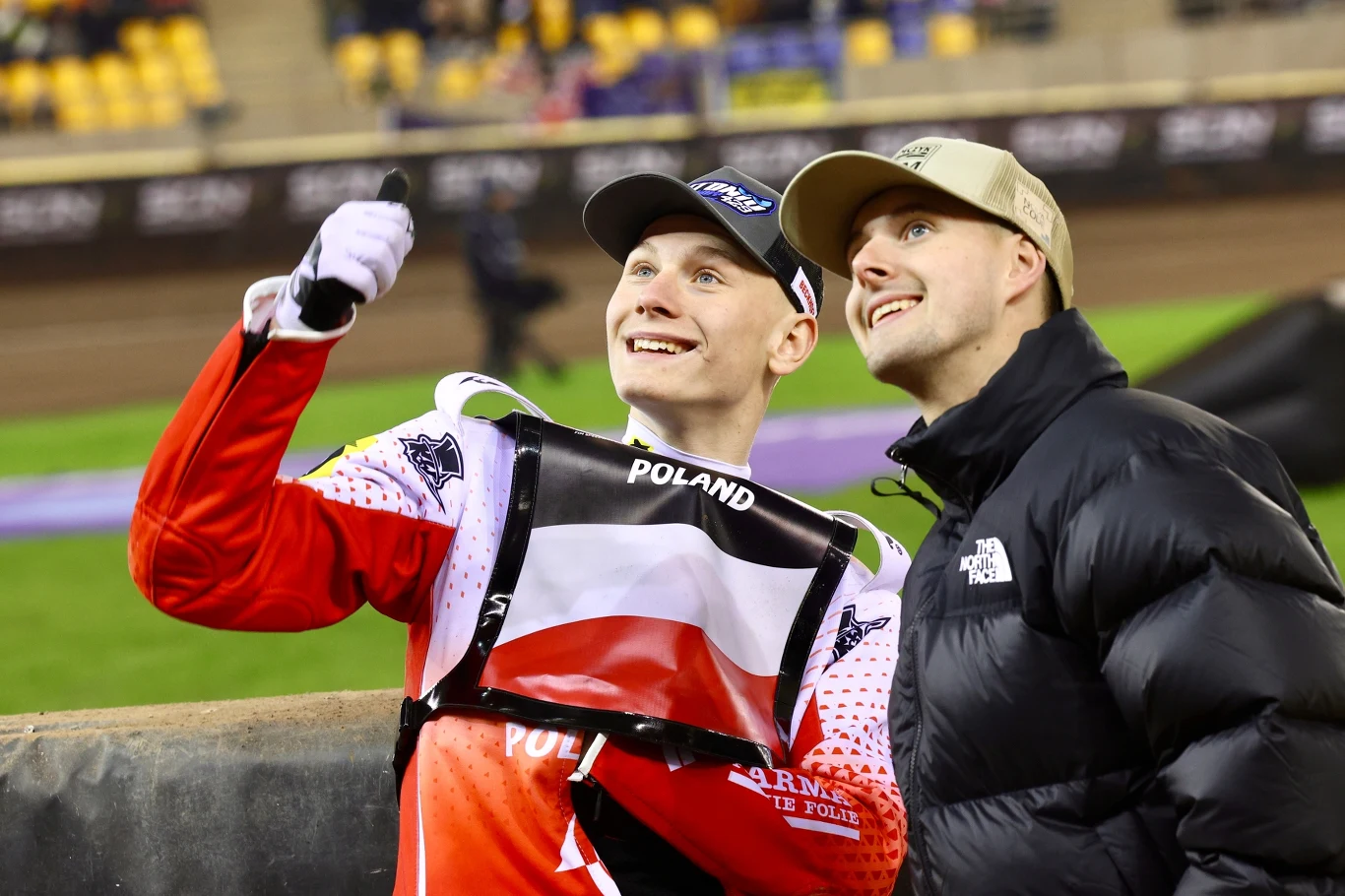 Dwóch młodych mężczyzn uśmiecha się i robi wspólne selfie na stadionie żużlowym, jeden ubrany w strój sportowy z polską flagą, drugi w czarnej kurtce i beżowej czapce.