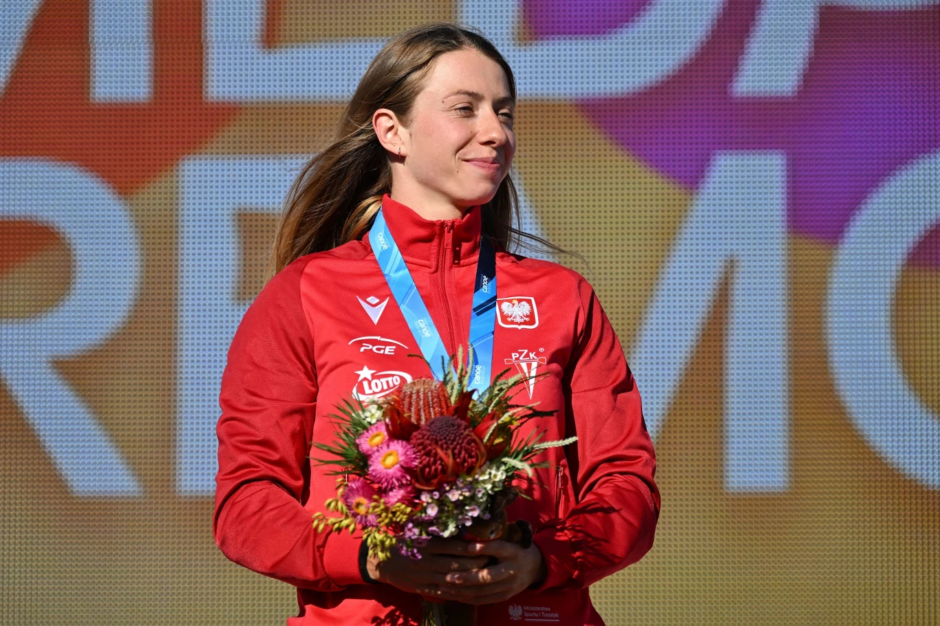 Sportowiec w czerwonym dresie reprezentacji Polski stoi na tle kolorowej planszy z medalem na szyi i bukietem kwiatów w dłoniach