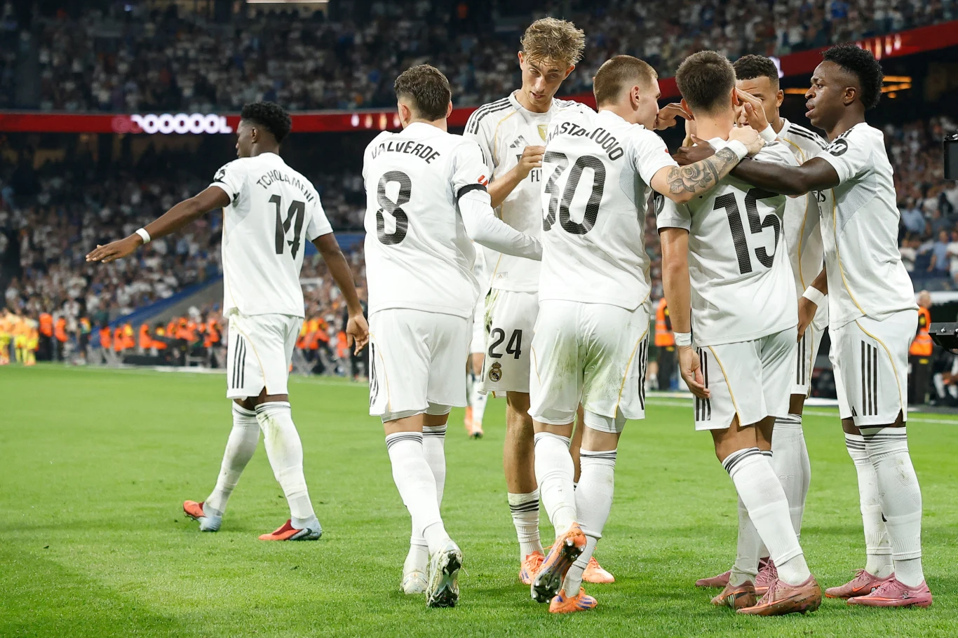 Grupa piłkarzy w białych strojach świętuje zdobycie gola na murawie stadionu podczas meczu przy pełnych trybunach kibiców.