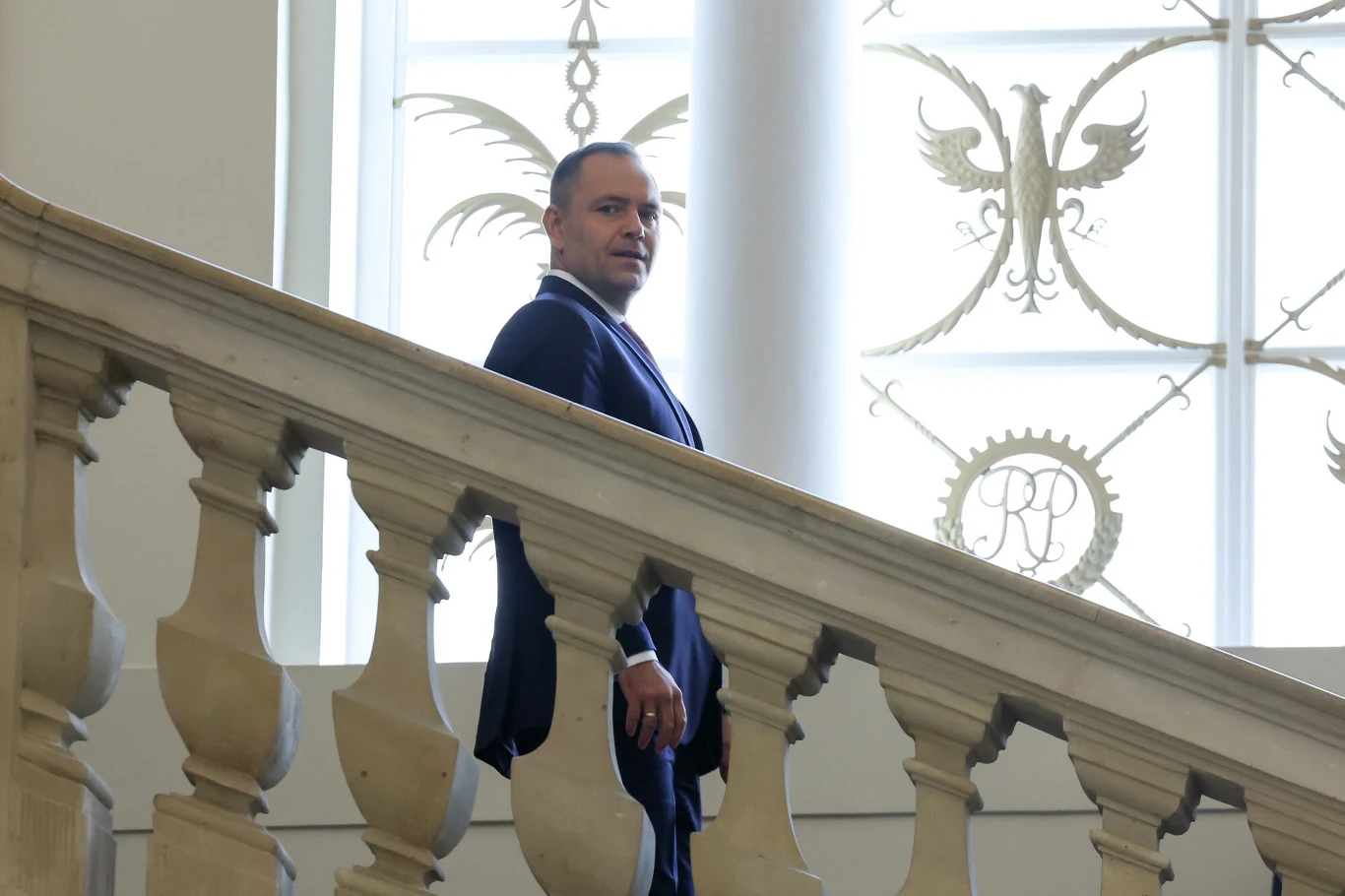 Elegancki mężczyzna w garniturze stoi na marmurowych schodach z klasyczną balustradą, w tle ozdobne okno z motywami orła i inicjałami.