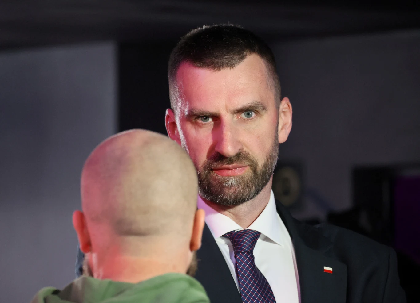 Mężczyzna w garniturze i krawacie, z poważnym wyrazem twarzy, patrzy na drugą osobę stojącą tyłem do obiektywu, która ma ogoloną głowę.