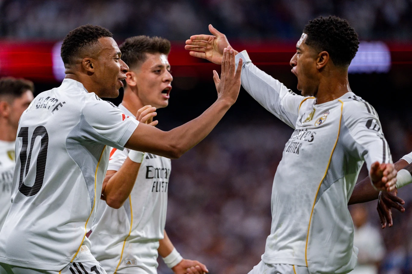 Piłkarze w białych strojach cieszą się po zdobyciu gola, wykonując energiczne gesty z wyciągniętymi ramionami na stadionie pełnym kibiców.