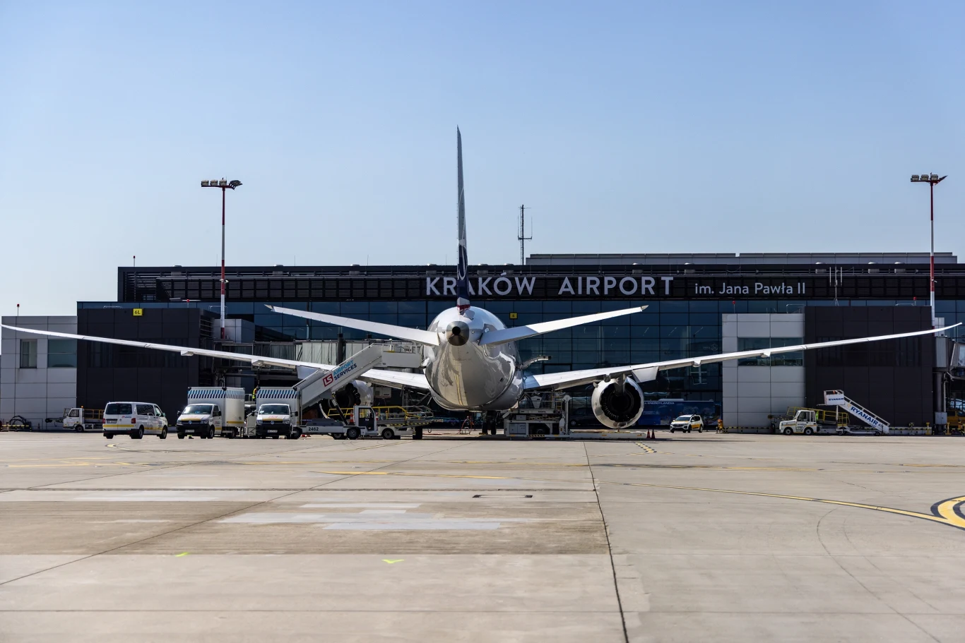 Samolot pasażerski stojący na płycie lotniska przed terminalem Kraków Airport im. Jana Pawła II w słoneczny dzień, wokół znajdują się pojazdy lotniskowe oraz budynek terminala z widocznymi napisami.