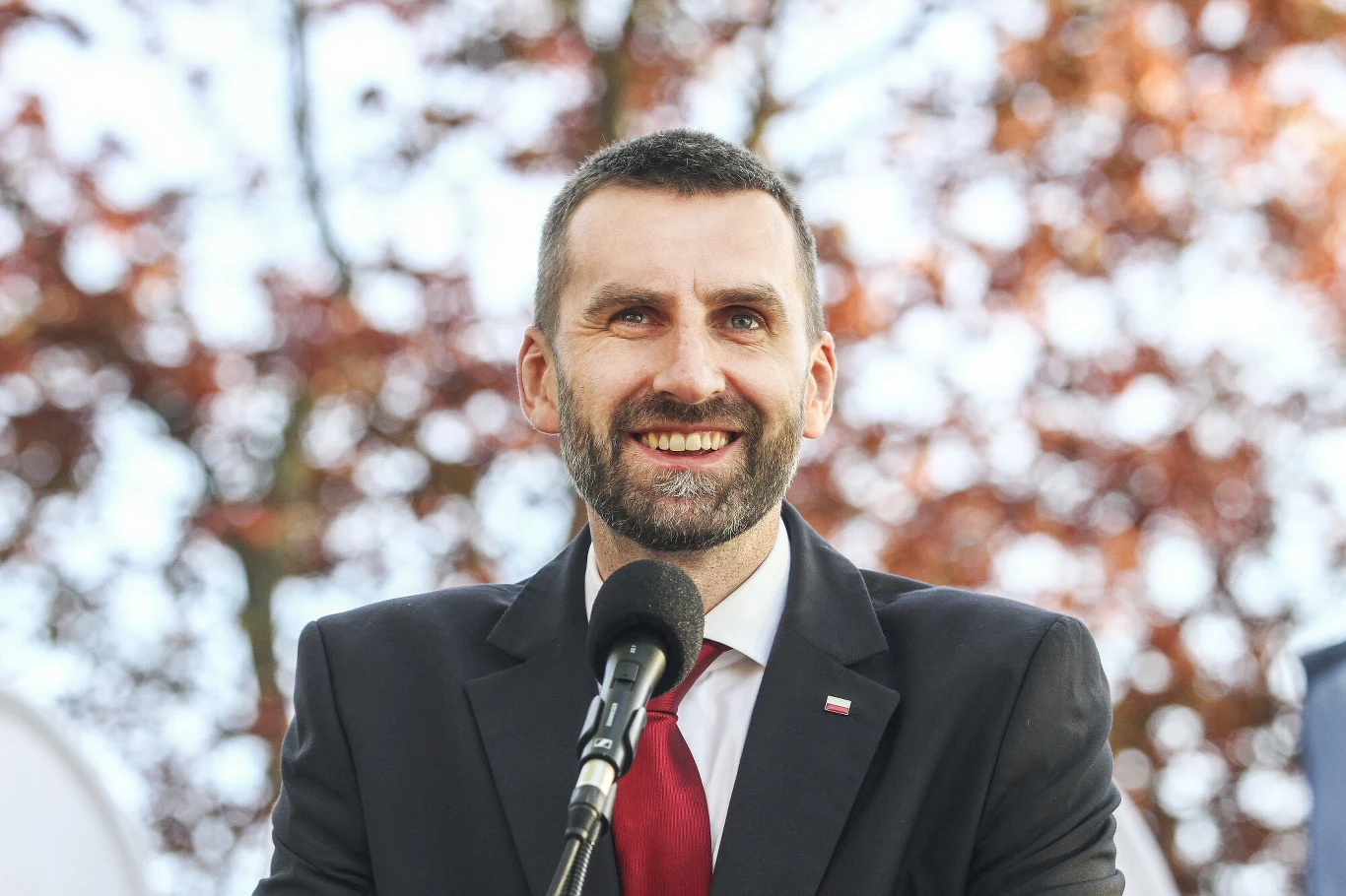 Mężczyzna w garniturze i czerwonym krawacie stoi przy mikrofonie podczas wystąpienia na świeżym powietrzu, w tle rozmyte jesienne liście.