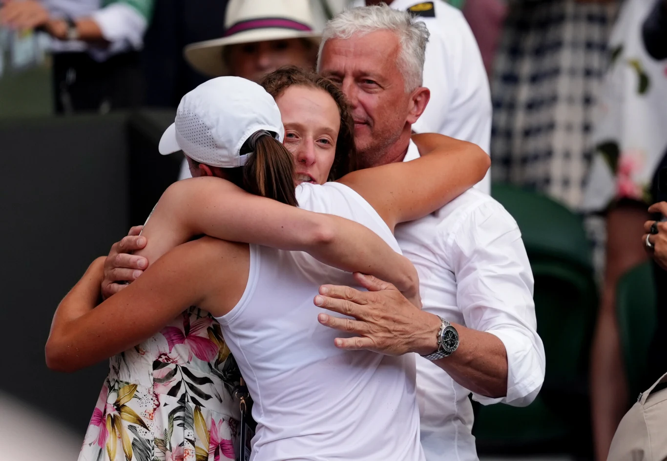 Grupa trzech osób w czułym uścisku, z których jedna ubrana jest w białą koszulkę i czapkę, a druga w sukienkę w kwiaty. W tle widoczne rozmazane postacie i fragment trybun.