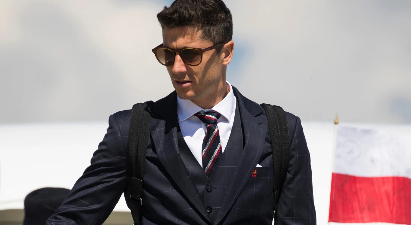 Elegancko ubrany mężczyzna w garniturze i okularach przeciwsłonecznych z plecakiem na ramionach, w tle flaga Polski i fragment samolotu.