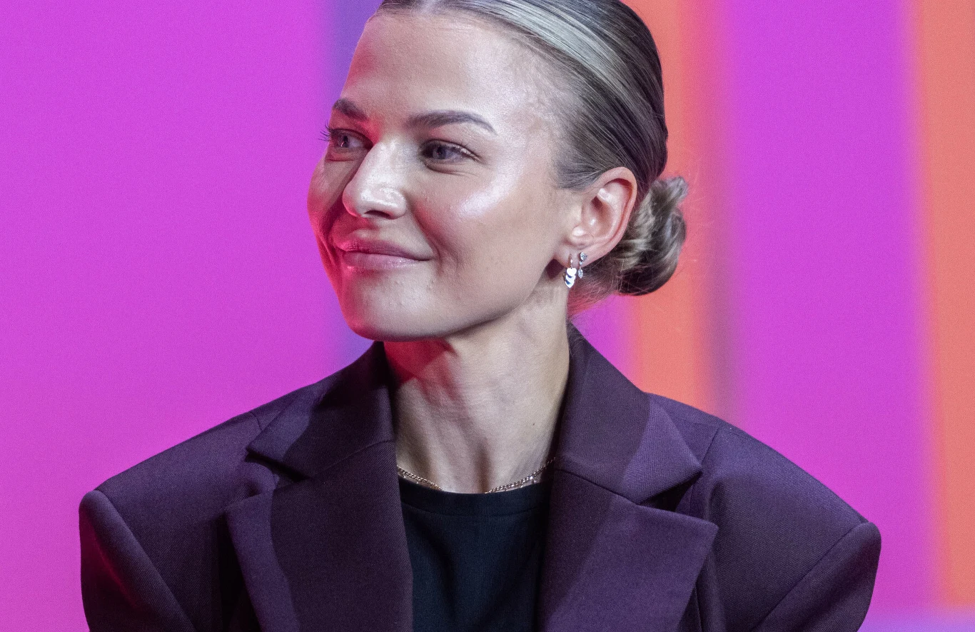 Kobieta z upiętymi włosami ubrana w fioletową marynarkę i czarną bluzkę, delikatny makijaż oraz niewielkie kolczyki, jednolite różowo-fioletowe tło.