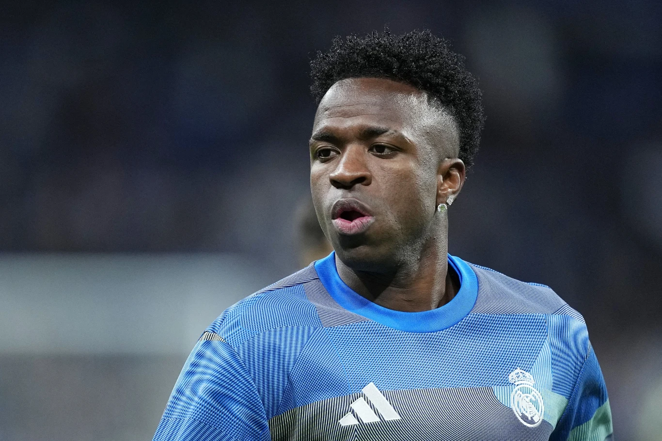 Młody mężczyzna ubrany w sportową koszulkę z logo klubu Real Madryt podczas meczu piłkarskiego, wyraz twarzy skupiony, tło rozmyte i ciemne.