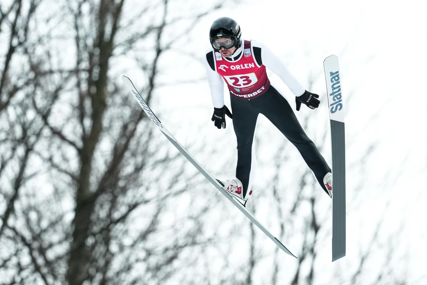 Sportowiec w trakcie skoku narciarskiego, ubrany w strój sportowy, kask oraz gogle, unosi się w powietrzu z rozpostartymi nartami, w tle rozmyte drzewa.