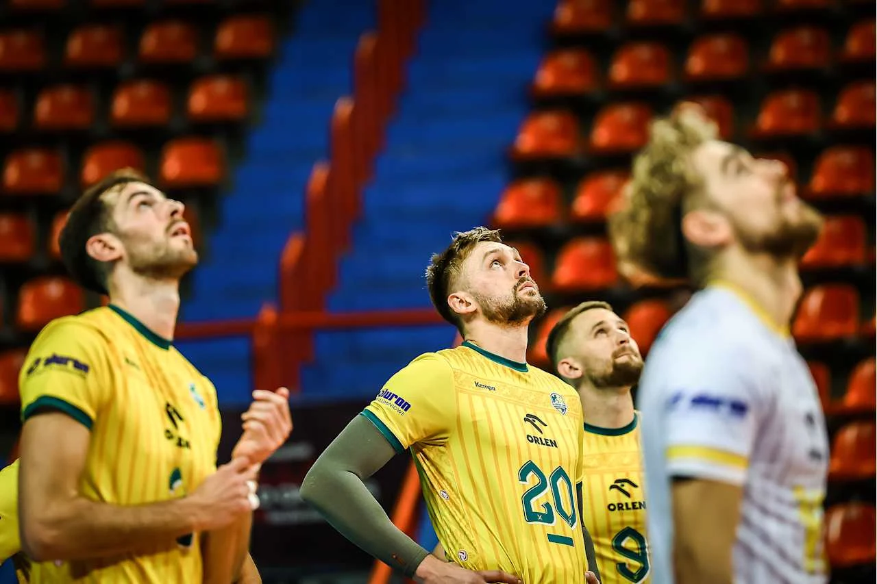 Grupa siatkarzy ubranych w żółte stroje z widocznymi numerami i logo sponsora patrzy w górę na tle pustych, pomarańczowych trybun.