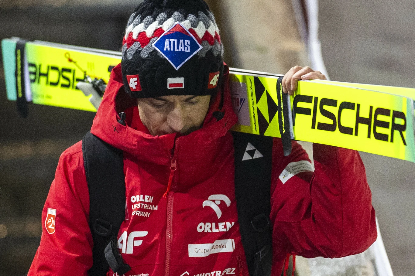 Mężczyzna ubrany w czerwoną kurtkę z licznymi logotypami sponsorów oraz czapkę w barwach czerwono-czarno-białych niesie na ramieniu narty z widocznym napisem Fischer. Postać wygląda na zamyśloną, głowę trzyma lekko pochyloną.