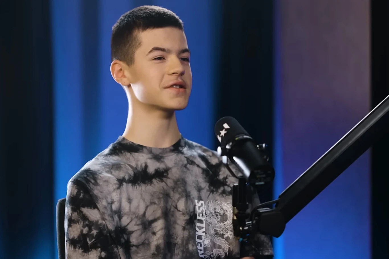 Młody mężczyzna w jasnej bluzie z czarno-szarym wzorem siedzi przed profesjonalnym mikrofonem studyjnym. Tło utrzymane w niebieskich odcieniach, sugerujące studio nagrań lub podcast.