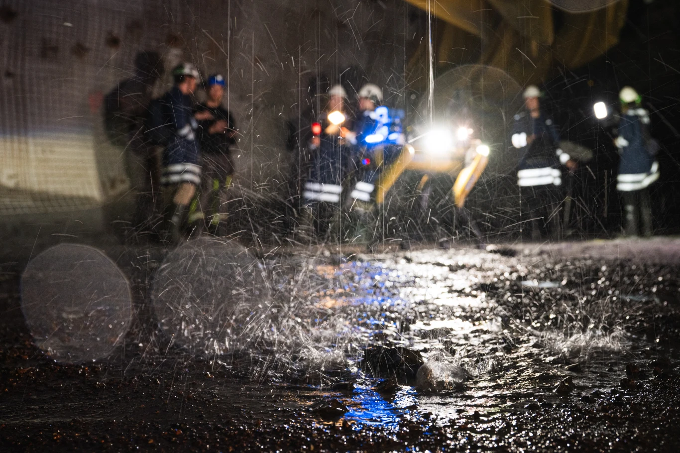 Podziemne tunele kopalni rudy żelaza LKAB w Kirunie Strażacy w odblaskowych strojach stojący w tle podczas intensywnych opadów deszczu, na pierwszym planie widoczna ulewa i spływająca po ziemi woda, w tle światła pojazdów ratowniczych.