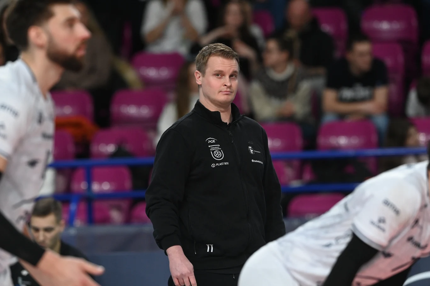 Trener w czarnej kurtce sportowej skupiony na boisku podczas meczu siatkówki, wokół rozmazani zawodnicy drużyny w białych koszulkach oraz różowe krzesełka na trybunach w tle.