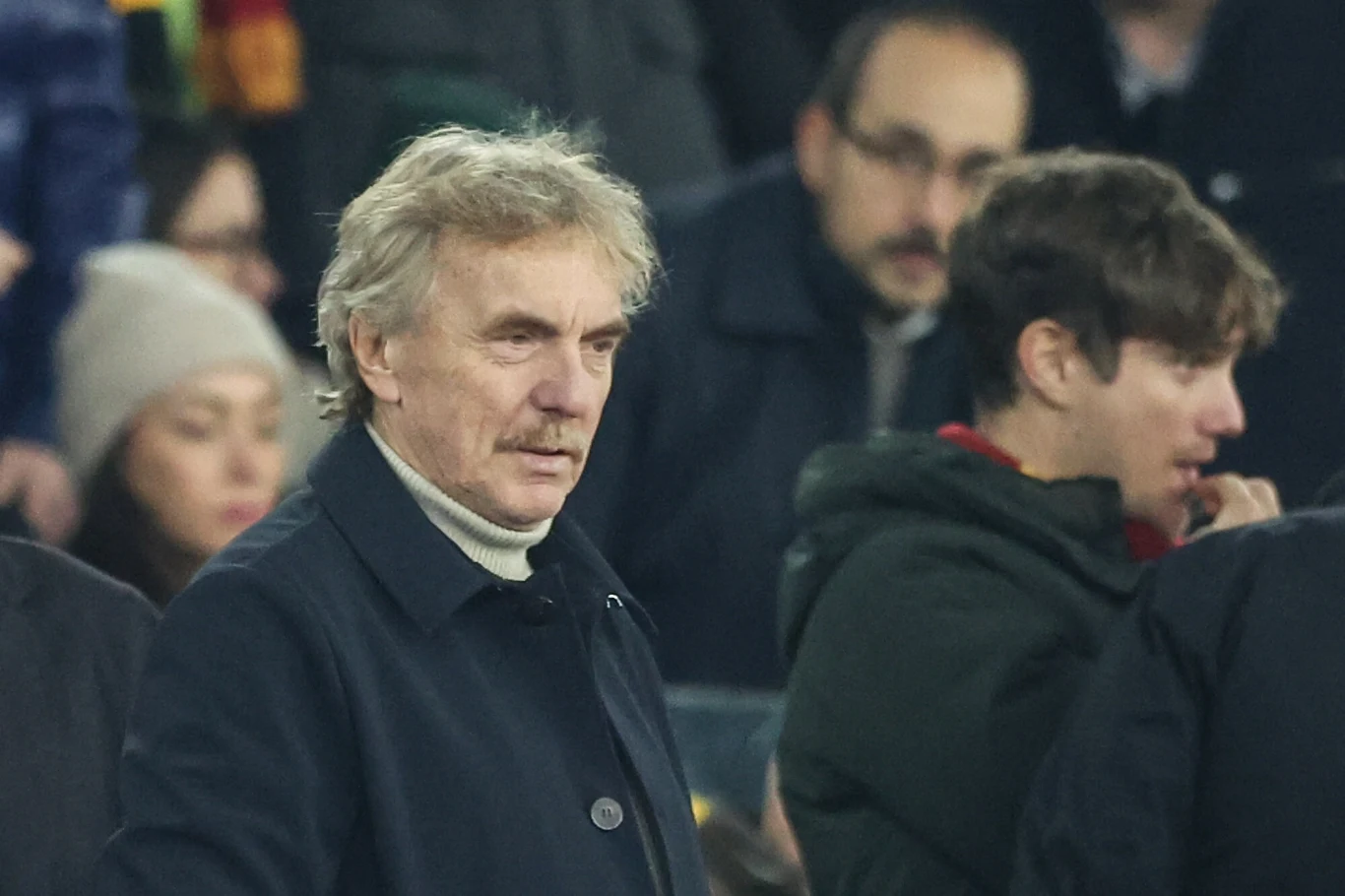 Mężczyzna w średnim wieku w jasnym golfie i ciemnym płaszczu stoi w otoczeniu ludzi na trybunach, wokół skupieni widzowie.