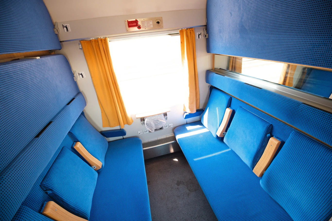 Wnętrze przedziału pociągu pasażerskiego z niebieskimi siedzeniami, żółtymi zasłonami przy oknie i jasnym światłem słonecznym przenikającym do środka.