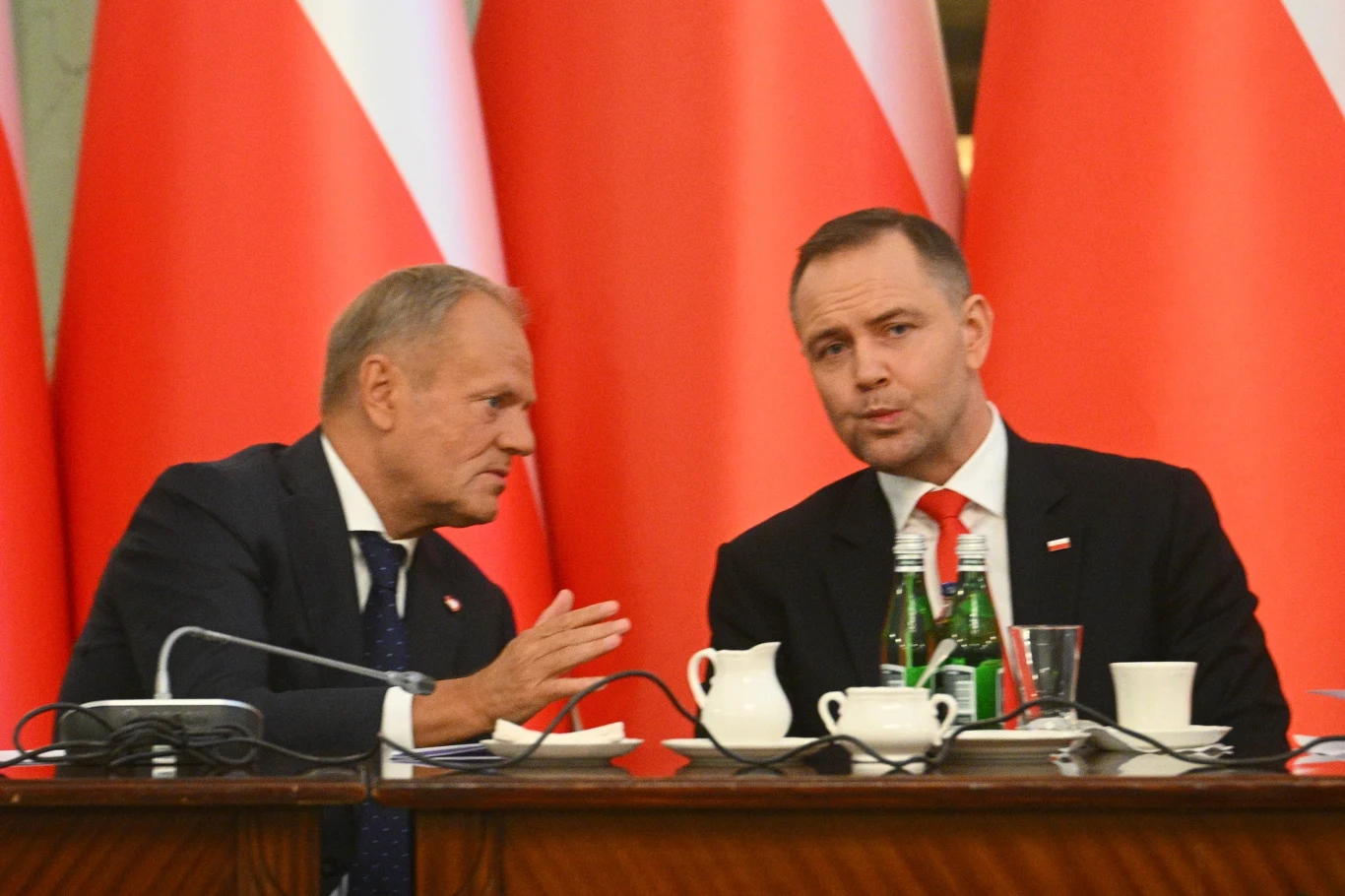 Donald Tusk i Karol Nawrocki Dwóch mężczyzn w garniturach siedzi przy stole, rozmawiają ze sobą. W tle widoczne duże biało-czerwone flagi. Na stole znajdują się butelki z wodą, filiżanki i dzbanek.