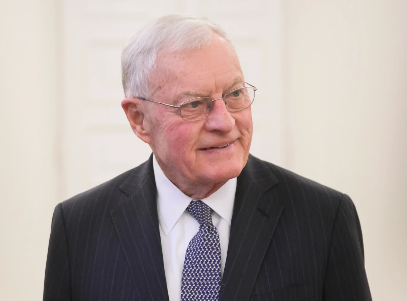 Starszy mężczyzna z siwymi włosami w okularach, ubrany w ciemny garnitur, białą koszulę i wzorzystą krawat. Stoi w jasnym, neutralnym pomieszczeniu i uśmiecha się lekko.