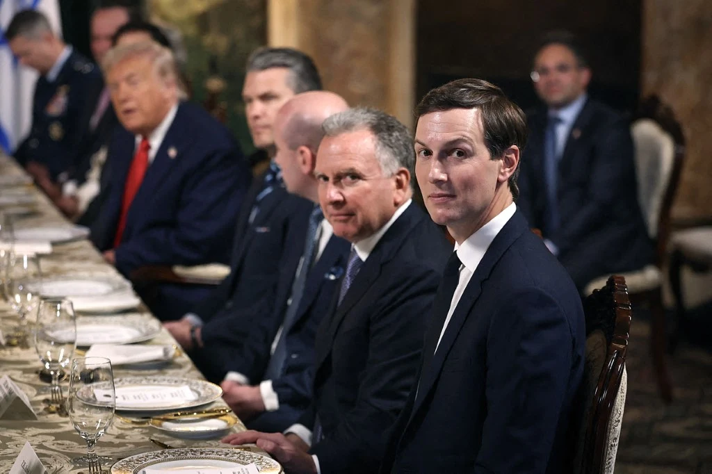 Grupa mężczyzn w garniturach siedzących przy elegancko nakrytym stole podczas oficjalnego spotkania lub kolacji. Na pierwszym planie młody mężczyzna zwrócony w stronę aparatu, w tle kilku innych uczestników spotkania.