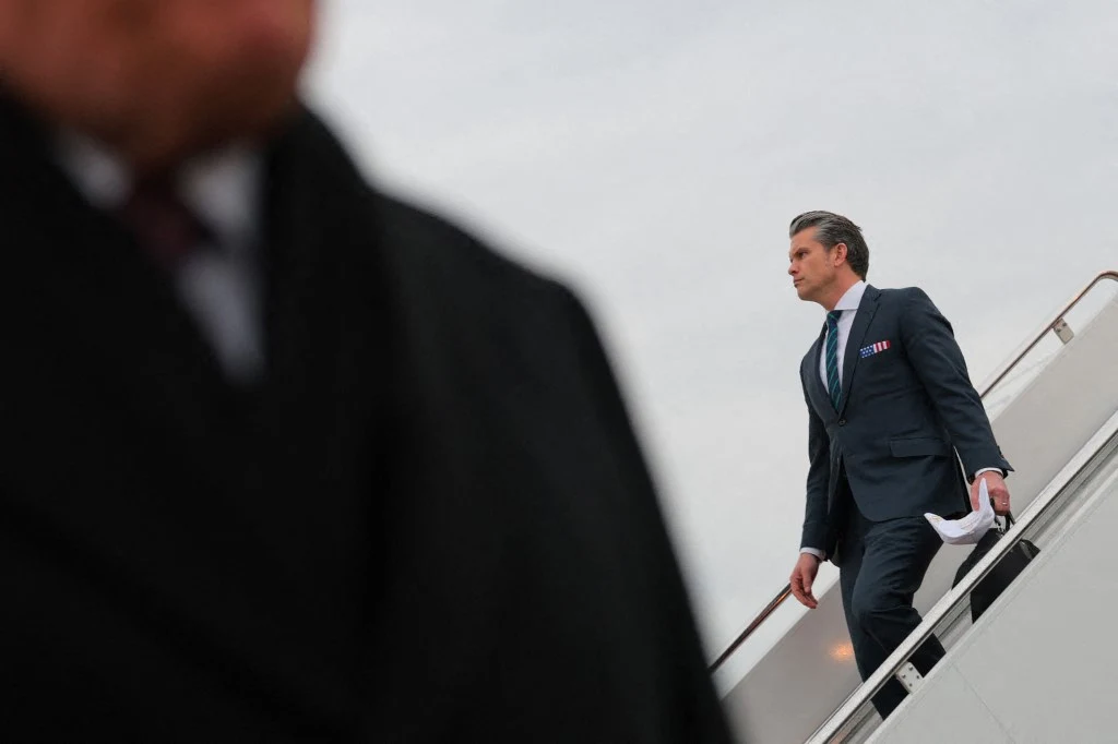 Mężczyzna w eleganckim garniturze trzymający dokumenty schodzi po schodach samolotu, na pierwszym planie częściowo widoczna sylwetka innej osoby w ciemnym płaszczu, nieostro przedstawionej.