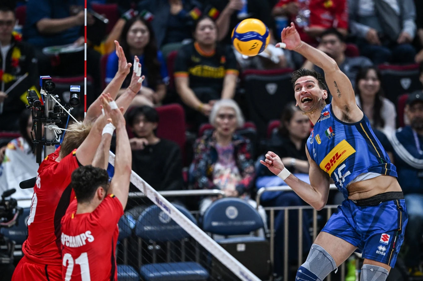 Yuri Romano to dwukrotny mistrz świata Siatkarz w niebieskim stroju wykonuje atak nad siatką, starając się zdobyć punkt, podczas gdy dwóch zawodników w czerwonych koszulkach próbuje zablokować piłkę. W tle widoczna publiczność uważnie obserwuje przebieg meczu.