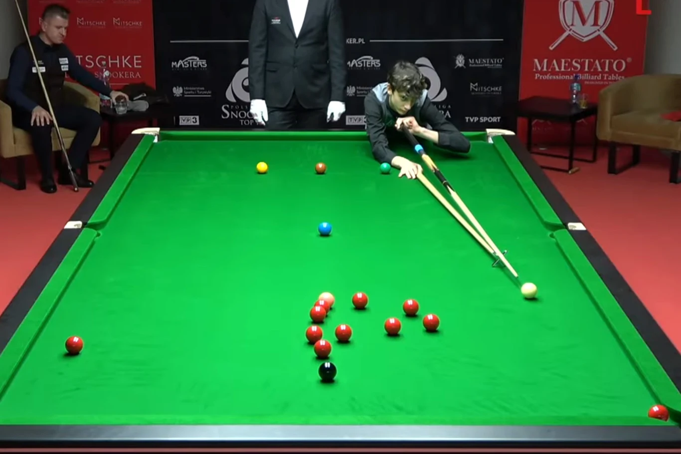 Zawodnik przy stole bilardowym wykonuje precyzyjne uderzenie kijem bilardowym przy użyciu specjalnej podpórki, na stole rozmieszczone są bile snookerowe, w tle sędzia oraz drugi zawodnik obserwujący rozgrywkę.
