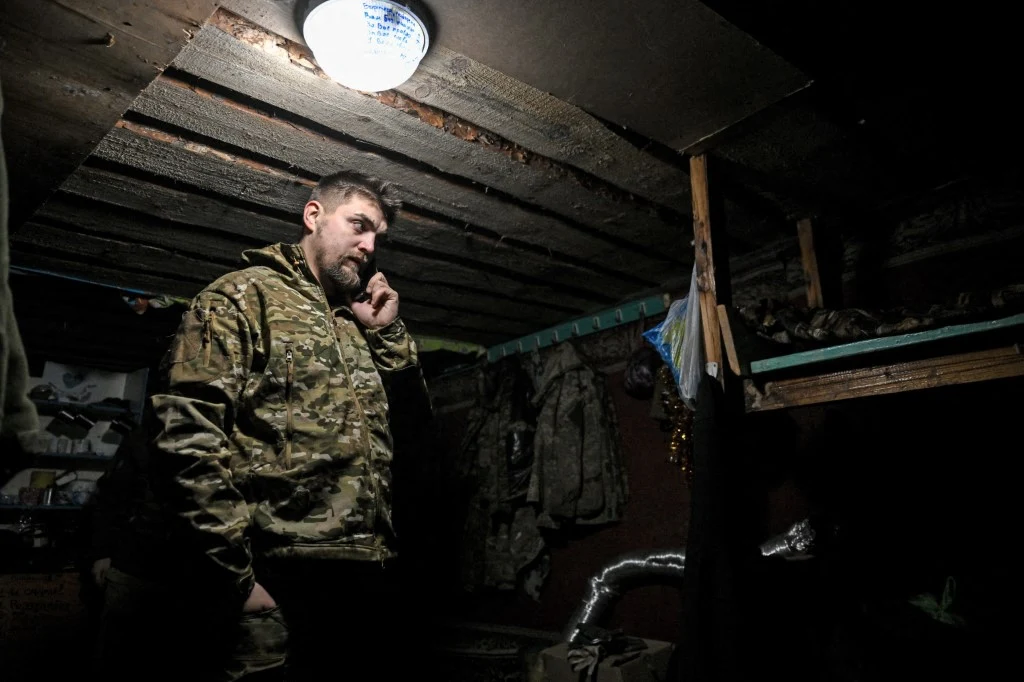 Mężczyzna w mundurze wojskowym stoi wewnątrz surowego, słabo oświetlonego pomieszczenia, trzymając przy uchu telefon. W tle widoczne powieszone na ścianie ubrania i łóżko piętrowe.