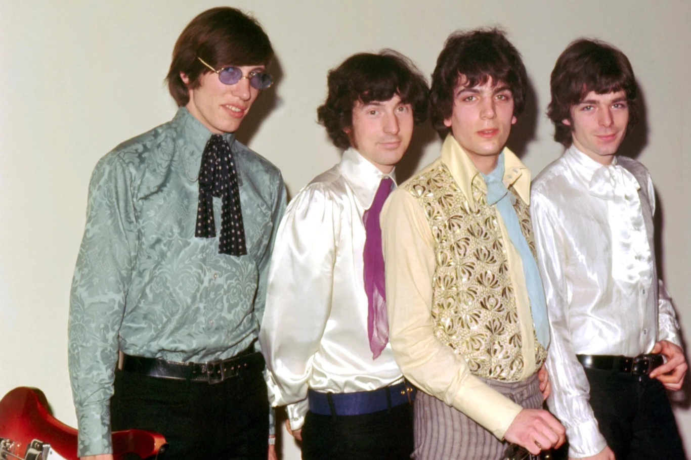 Czterech młodych mężczyzn ubranych w kolorowe, eleganckie koszule i krawaty, stojących obok siebie na jasnym tle; jeden z nich trzyma gitarę.