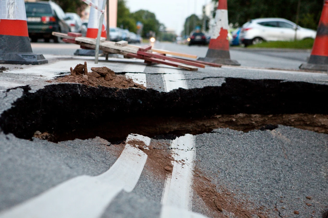 Duża wyrwa w asfalcie na ulicy otoczona pachołkami ostrzegawczymi i barierkami zabezpieczającymi ruch pojazdów. Linie jezdni przerywane w miejscu zapadnięcia się nawierzchni, na dalszym planie zaparkowane samochody i drzewa.