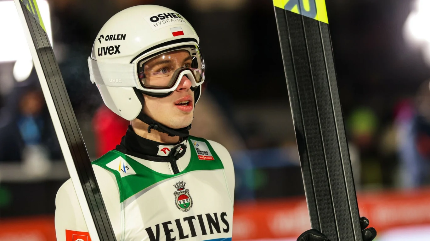 Skoczek narciarski w kombinezonie sportowym oraz kasku, trzymający narty, stoi na tle rozmytego stadionu z widocznymi światłami.