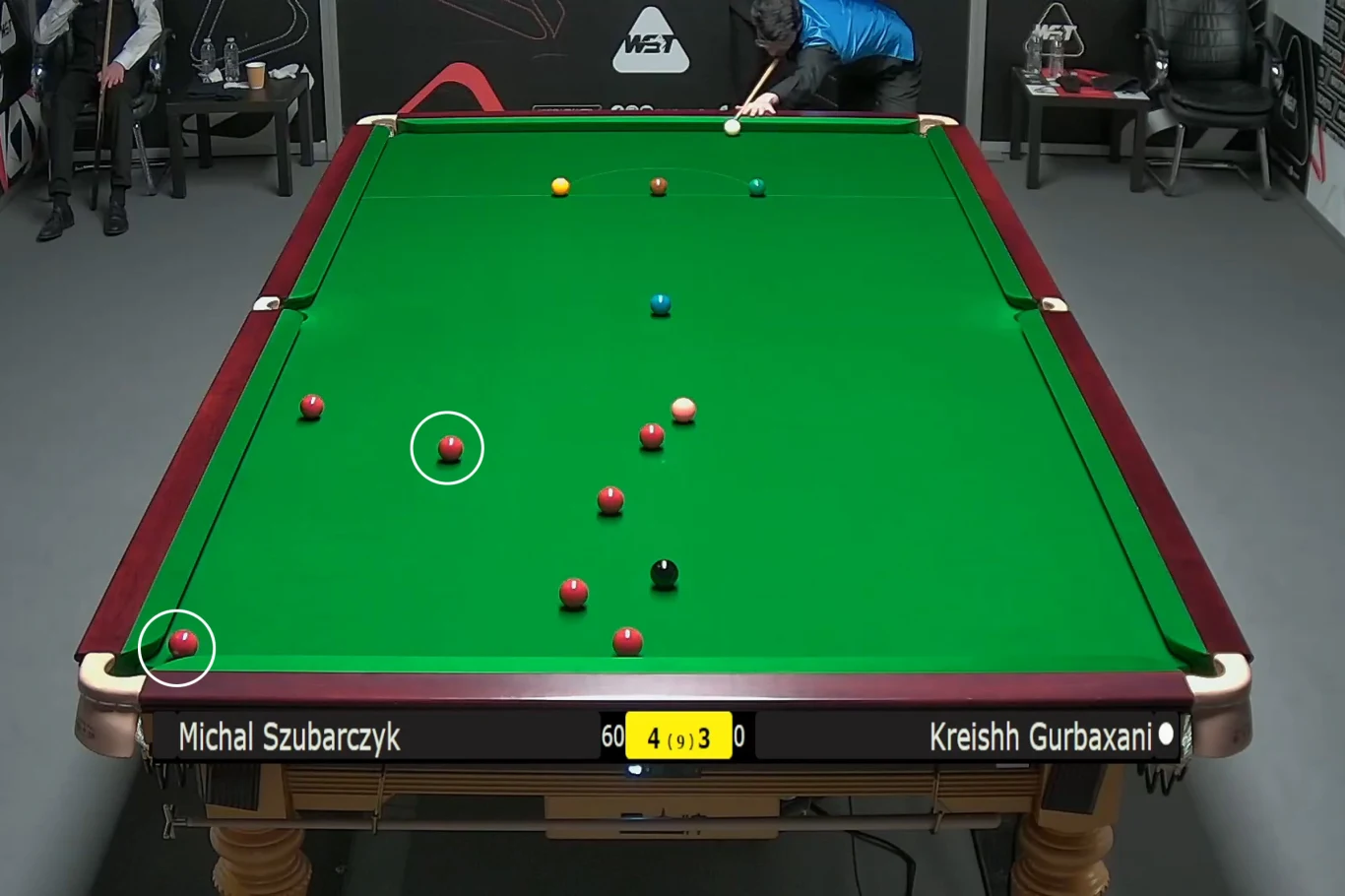Stół bilardowy do snookera z rozłożonymi kolorowymi bilami, dwóch graczy skoncentrowanych nad stołem, nazwiska graczy oraz aktualny wynik wyświetlone na tablicy wyników.