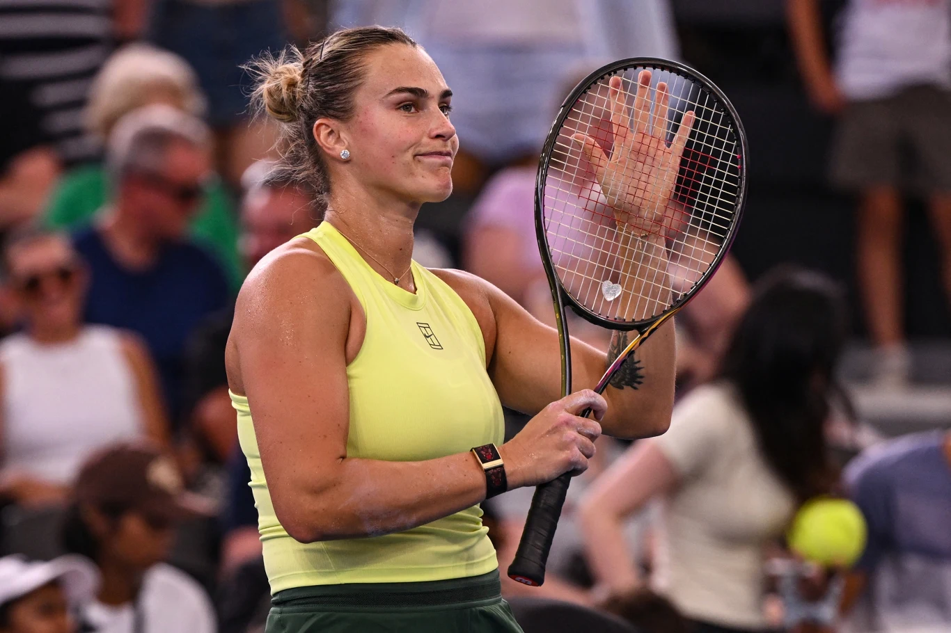 Tenisistka w żółtym stroju trzymająca rakietę tenisową, unosząca rękę w geście pożegnania lub podziękowania na tle rozmytej publiczności na trybunach.