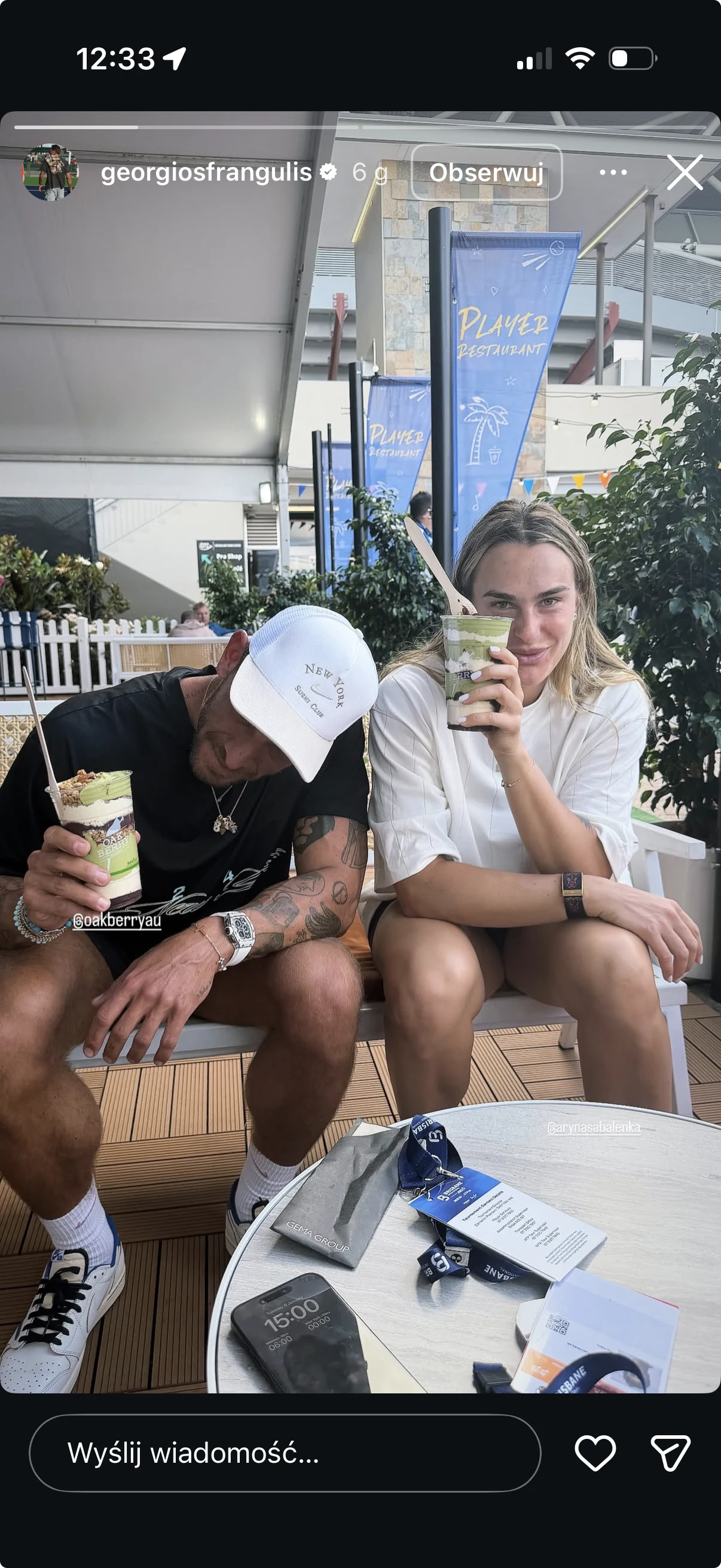 Georgios Frangulis z Aryną Sabalenką Dwie osoby siedzą przy okrągłym stoliku na świeżym powietrzu, trzymają napoje w plastikowych kubkach, jedna z nich nosi białą czapkę z daszkiem i czarny T-shirt, druga ma jasne ubranie i rozpuszczone blond włosy, na stole leżą przedmioty codziennego uż...