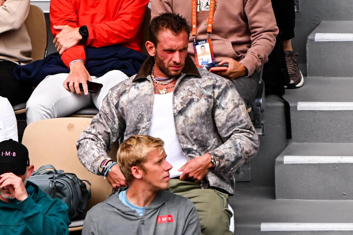 Georgios Frangulis Mężczyzna w kamuflażowej kurtce siedzi na trybunach obok innych widzów, trzyma rękę przy pasku spodni. W tle widoczne inne osoby, jedna z identyfikatorem na szyi. Atmosfera trybun sportowego wydarzenia.