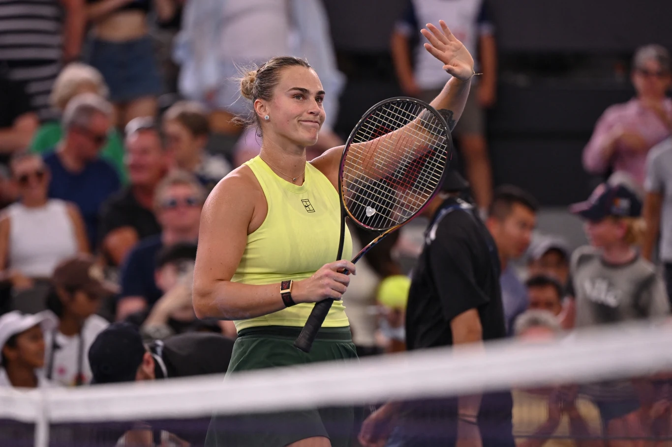 Aryna Sabalenka Tenisistka w sportowym stroju, trzymająca rakietę, macha ręką w stronę publiczności na korcie, w tle widzowie na trybunach.