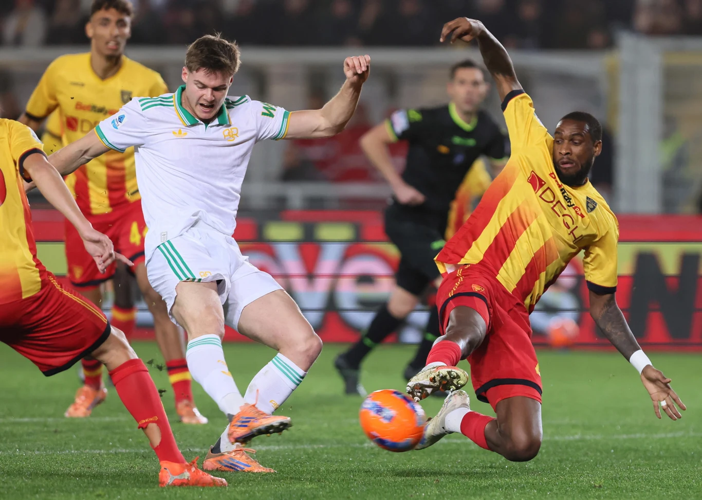 Piłkarze dwóch drużyn rywalizują o piłkę podczas meczu, zawodnik w żółto-czerwonym stroju wykonuje wślizg próbując odebrać piłkę rywalowi w białym stroju, wokół nich widoczni są inni zawodnicy oraz sędzia.