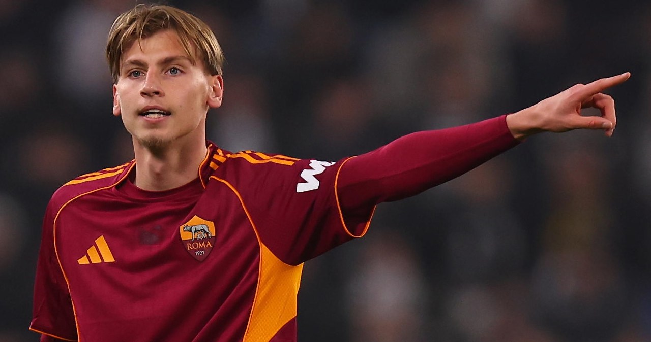 Piłkarze dwóch drużyn rywalizują o piłkę podczas meczu, zawodnik w żółto-czerwonym stroju wykonuje wślizg próbując odebrać piłkę rywalowi w białym stroju, wokół nich widoczni są inni zawodnicy oraz sędzia.