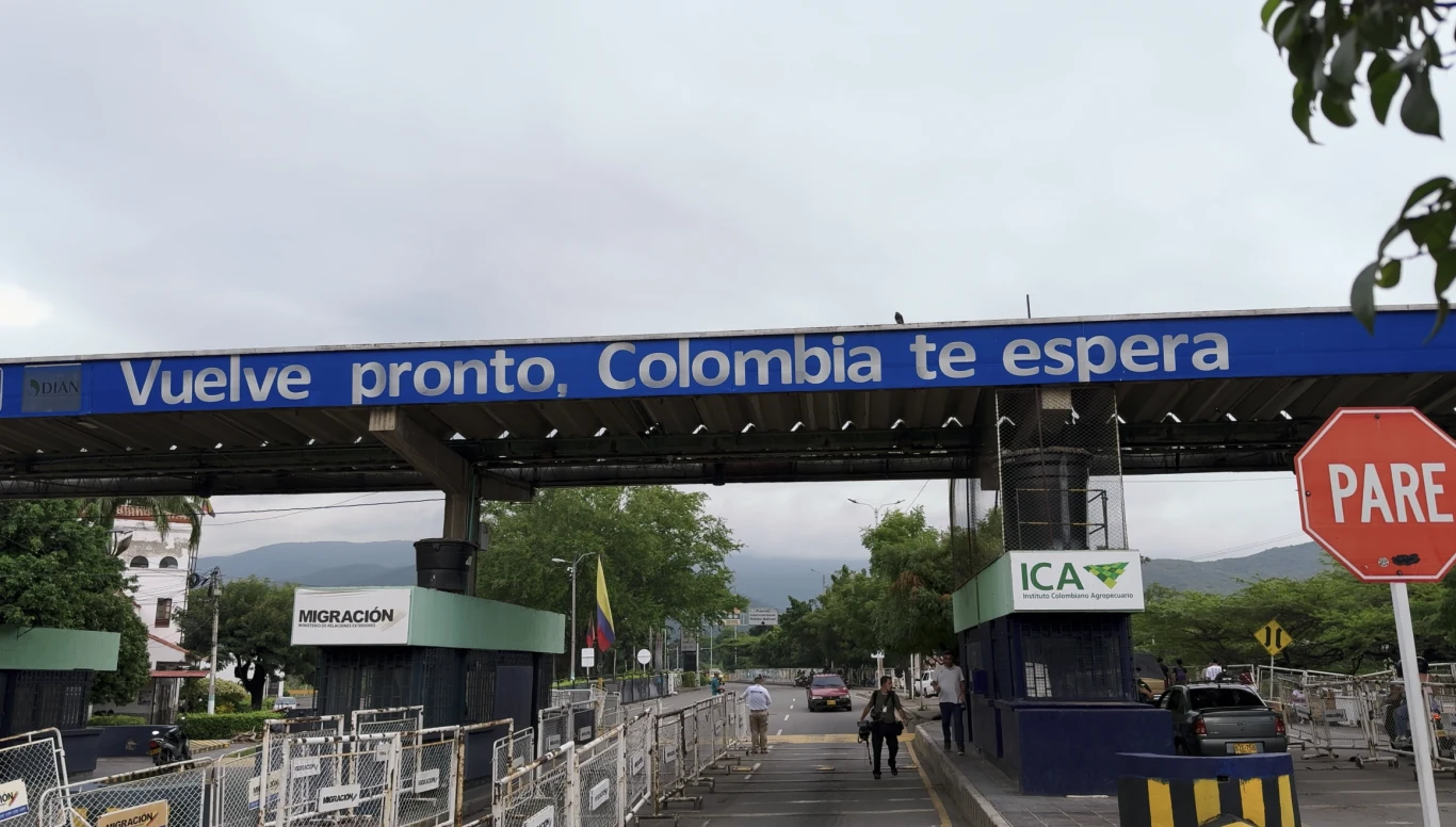 Przejście graniczne z napisem powitalnym, po lewej widoczny budynek służby imigracyjnej, po prawej stanowisko kontroli ICA, znak stop oraz kilka osób przechodzących przez granicę; w tle zieleń i góry.