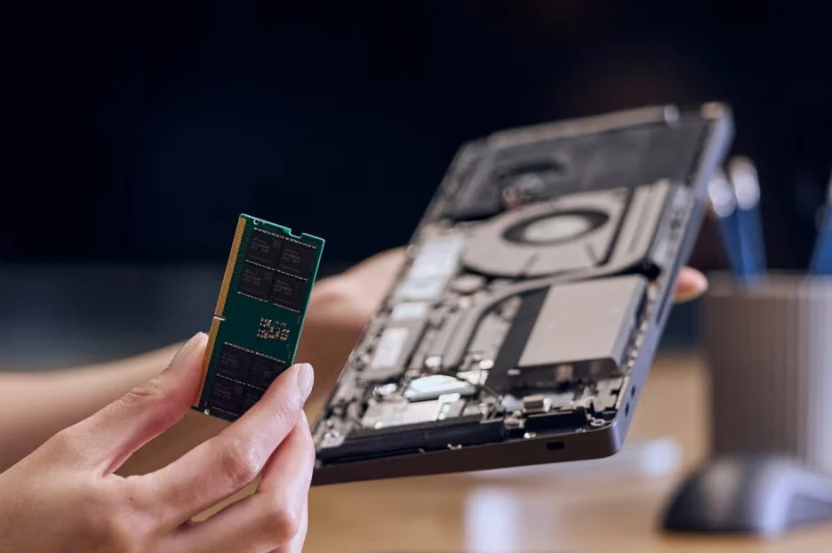 Osoba trzyma wyjętą pamięć RAM oraz otwartego laptopa pokazującego wnętrze z widoczną płytą główną i podzespołami elektronicznymi.