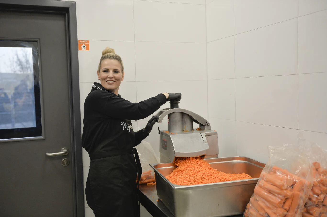 Kobieta ubrana w fartuch obsługuje maszynę do rozdrabniania marchewek, wsypując starte warzywa do dużego stalowego pojemnika; obok znajdują się worki z marchewkami w całości, a tło stanowi jasna kafelkowana ściana.