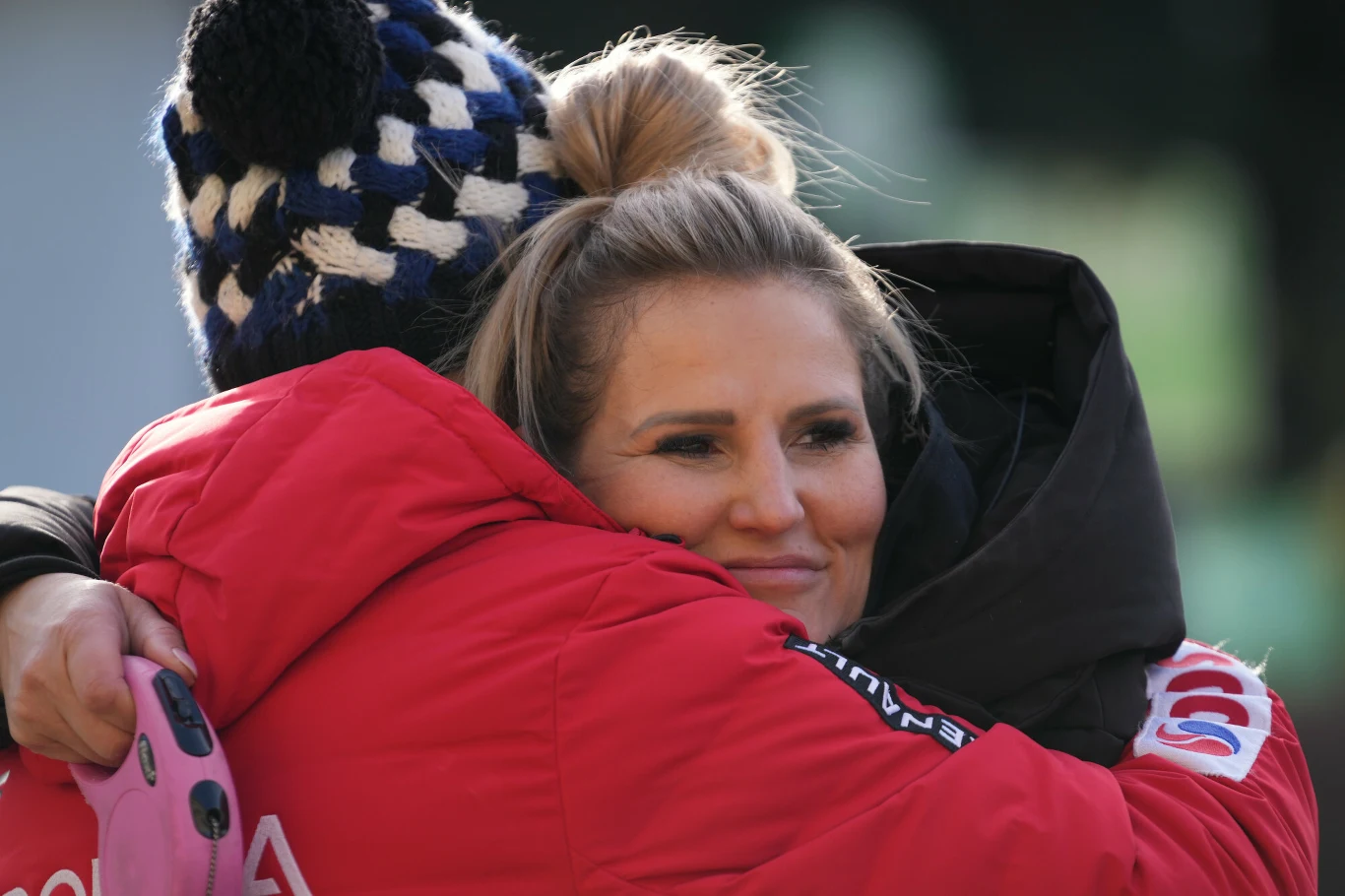 Dwie osoby obejmujące się, jedna z nich uśmiechnięta, ubrana w ciepłą kurtkę, druga ma na sobie czapkę z czarno-białym wzorem.