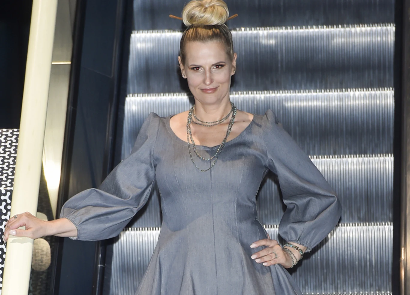 Kobieta w eleganckiej, szarej sukience z balonowymi rękawami stoi na tle ruchomych schodów, z włosami upiętymi w kok oraz licznymi naszyjnikami i bransoletkami, prezentuje się w pewnej pozycji.