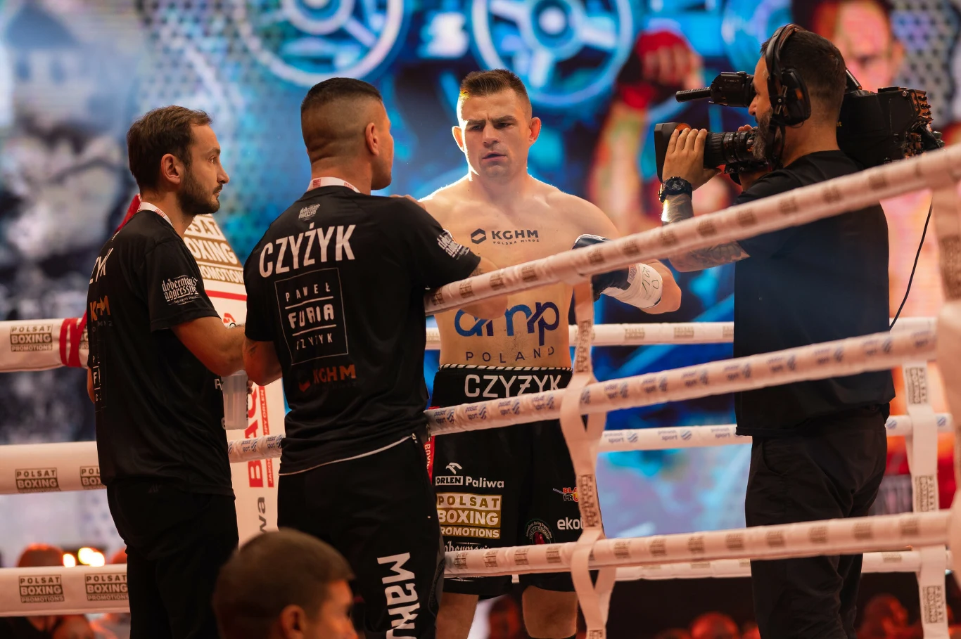 Bokser stojący w ringu w otoczeniu trenerów oraz kamerzysty, światła sceniczne i tło z grafiką sportową, na spodenkach widoczne logo sponsorów.