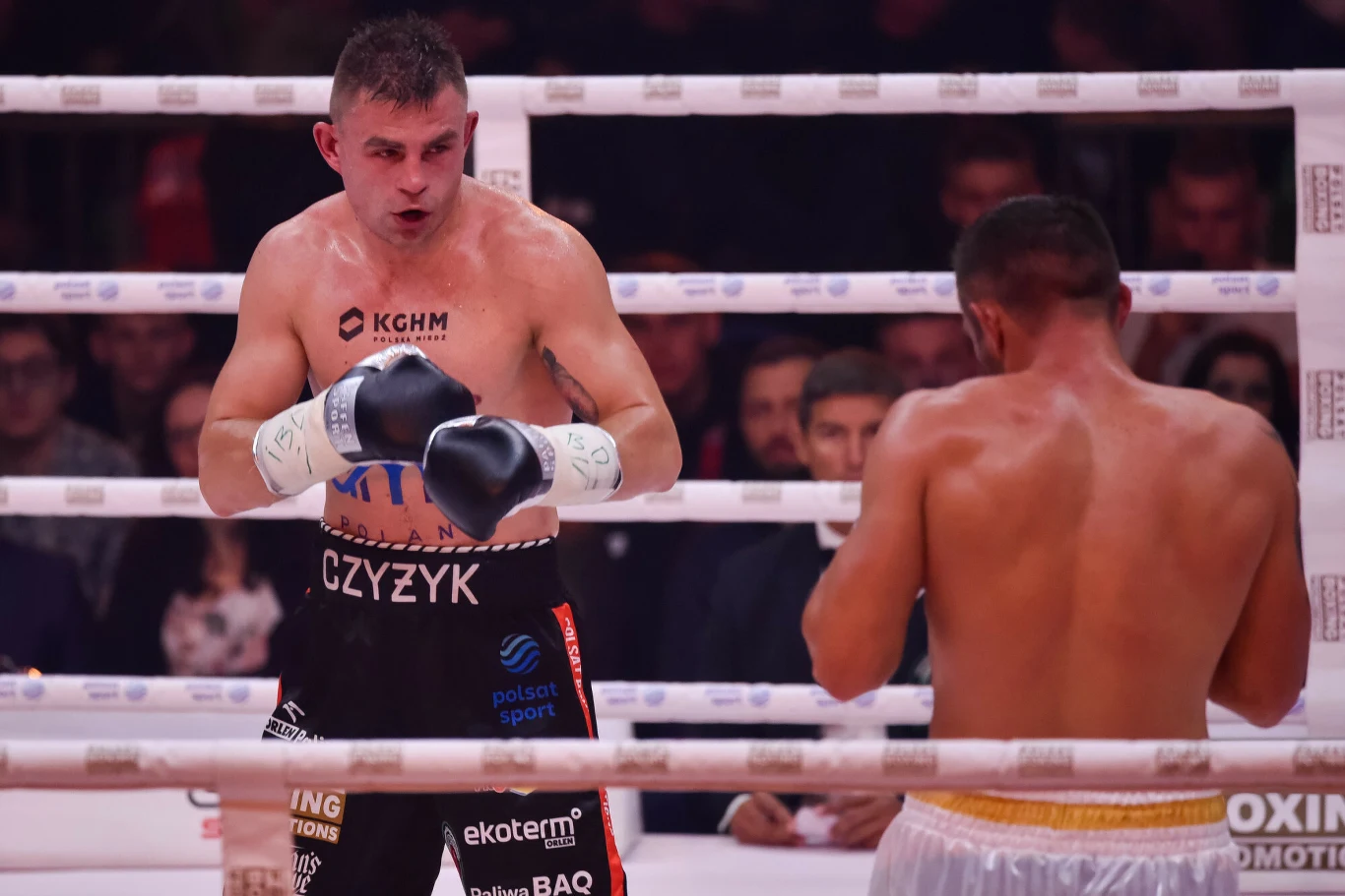 Dwóch bokserów walczy na ringu podczas pojedynku bokserskiego. Jeden zawodnik nosi rękawice i spodenki z nazwiskiem, drugi stoi naprzeciwko z rękawicami przygotowanymi do obrony. Tło wypełnione publicznością oraz jasno oświetlonym ringiem.