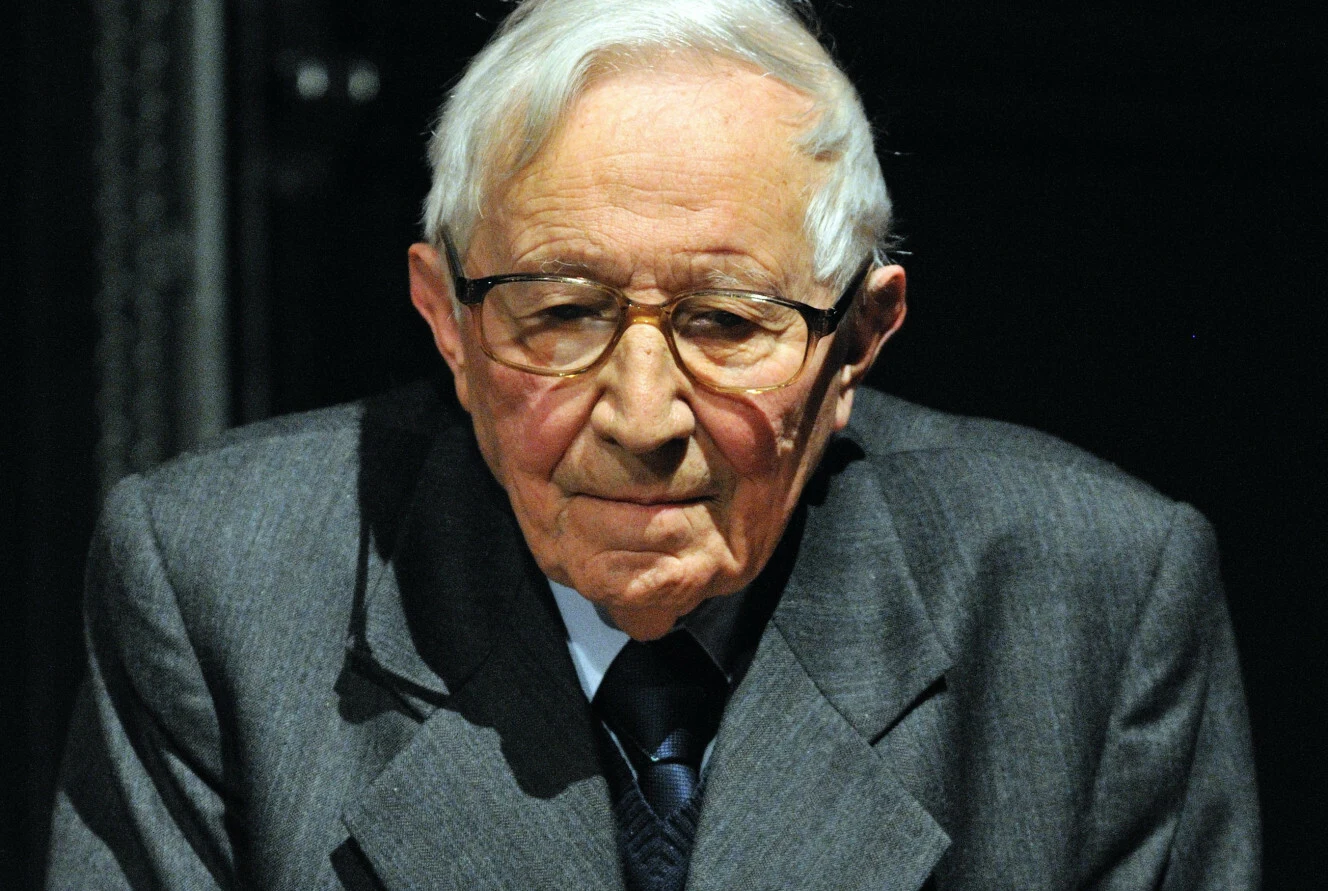 Starszy mężczyzna w okularach, ubrany w elegancki garnitur i krawat, patrzy spokojnie w stronę aparatu. Siwe włosy, poważny wyraz twarzy, jednolite ciemne tło przykuwa uwagę do postaci.